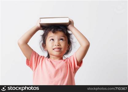 Asian little girl holding a book on head and eyes looking top on white background head. On face a cute girl so happy and smile.