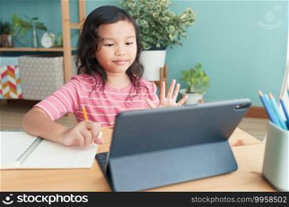 Asian little girl waving in greeting online tutor on tablet digital in interior at home morning. Concept of online learning at home