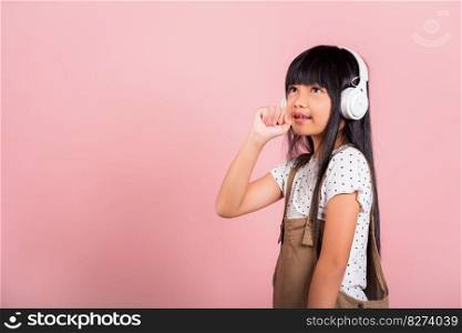 Asian little kid 10 years old smile listening music wear wireless headset and keeps hand near mouth sings song, studio shot isolated on pink background, Child girl funny listen music with headphones