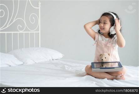 Asian little kid girl wear headphones listening music with happy face sitting on bed with tablet and hold bear doll at bedroom