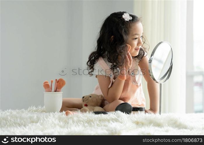 Asian little kid girls enjoy playing mother?s cosmetic make up on bed. Cute child uses a pink brush to color on the cheeks while looking in the mirror at bedroom
