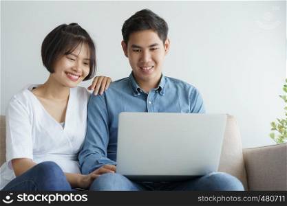 Asian love couple sitting on the sofa and using laptop they is smiling happily at home