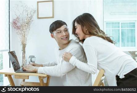 Asian lovely couple smiling and looking at each other while using laptop and working at home. Lifestyle, Valentine and Love Concept.