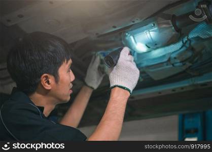 Asian male mechanical hold and shining flashlight to examine car under chassis of automotive vehicle. Safety suspension inspection check service maintenance for customer before road trip concept