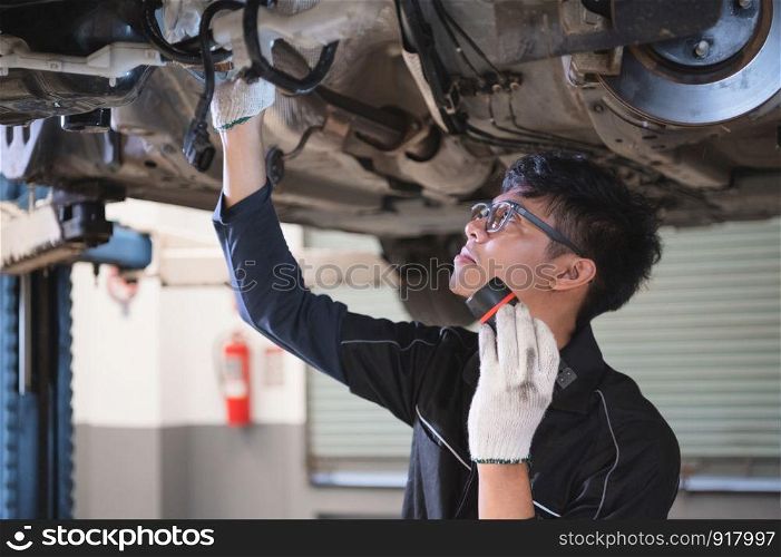 Asian male mechanical hold and shining flashlight to examine car under chassis of automotive vehicle. Safety suspension inspection check service maintenance for customer before road trip concept