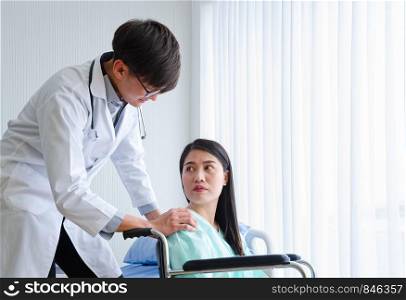Asian male Medicine Physician Doctor wearing glasses and encouraging disabled female patient sitting on wheelchair in white hospital room. Medical and Healthcare concept of patient paralysis loss human lifestyle