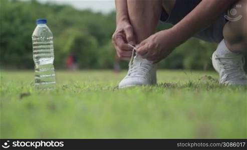Asian male sitting down tie shoes laces on green grass field during Exercise at the park with trees Background, outdoor exercising, active lifestyle, stay hydrated water bottle on the side, front view