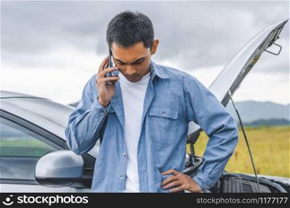 Asian man calling car maintenance service after having accident during travel long weekend between road trip. Car broken in mountain meadow background. People transportation. Need help insurance claim