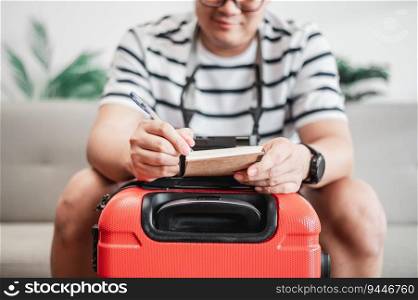 Asian man glasses is preparing clothes in suitcases. He’s choosing clothes, travel documents, the itinerary for a solo trip, and check the checklist. Travel, holiday and vacation concept.