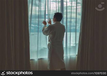 Asian man in bathrobe suit opening and sightseeing the sea beach when waking up from the bed in luxury hotel in morning, Life style and holiday concept