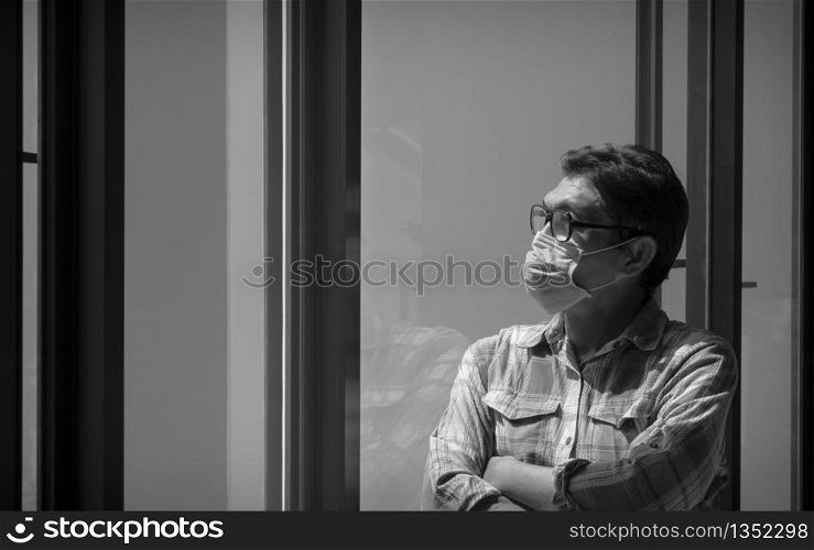 Asian man in plaid long sleeve shirt wearing protective mask and looking out of the glass window at home, He hopes that the spreading of the Coronavirus problem will end soon, black and white style