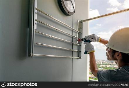Asian man screwing screw install clothesline with cordless drill. Technician wears safety helmet and hand holding drilling tool for apartment renovation. Man working home improvement with DIY concept.