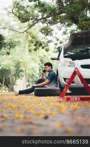 Asian man sitting beside car and using mobile phone calling for assistance after a car breakdown on street. Concept of vehicle engine problem or accident and emergency help from Professional mechanic