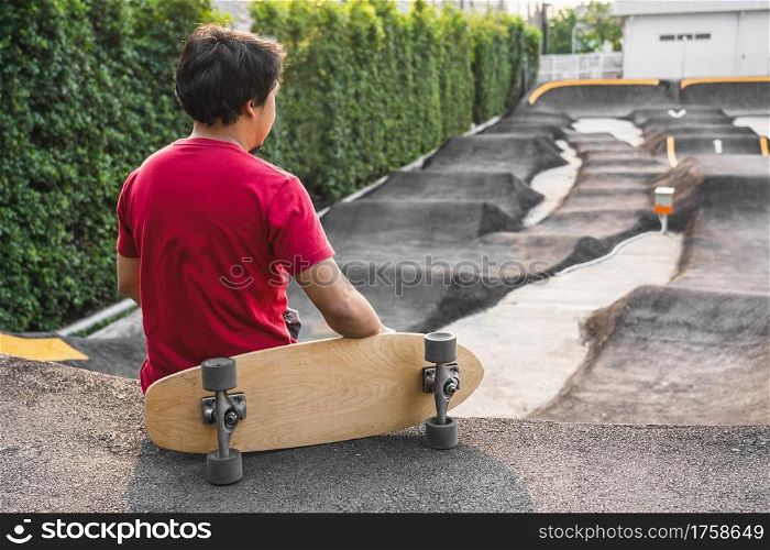 Asian man sitting with surfskate or skate board in pumptrack skate Park when sunrise time over photo blur of pumptrack curve, extream sport, healthy and exercise, fashion in covid19 concept