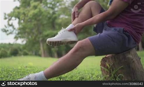Asian man twisted his ankle during exercise Inside the park sitting on tree trunk with lawn on the background, ankle pain, joint ligament problem, painful face expression, risk of accident