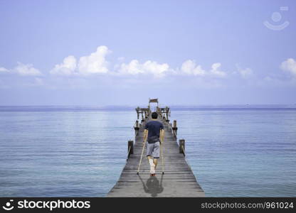Asian man used wooden crutches walks on bridge pier boat in the sea and the bright sky at Koh Kood, Trat in Thailand.
