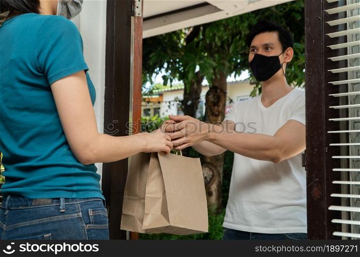 Asian man wearing a protective mask and delivering packages from grocery to young female customers at her home after online order. Concept of transportation service and express delivery and safety