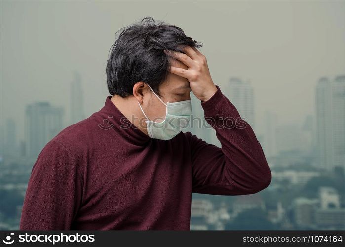 Asian man wearing the face mask against air pollution with hand catching the headache at the balcony of High Apartment which can see pollution and heavy fog over the bangkok cityscape background