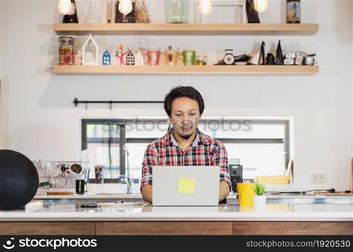 Asian man working from home with technology laptop and mobile phone at kitchen room, Working at home in Covid-19 pandemic, new normal concept