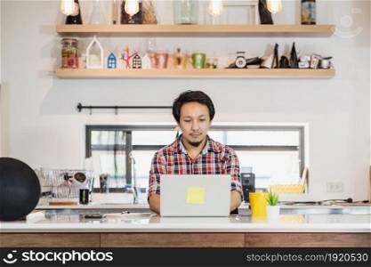 Asian man working from home with technology laptop and mobile phone at kitchen room, Working at home in Covid-19 pandemic, new normal concept