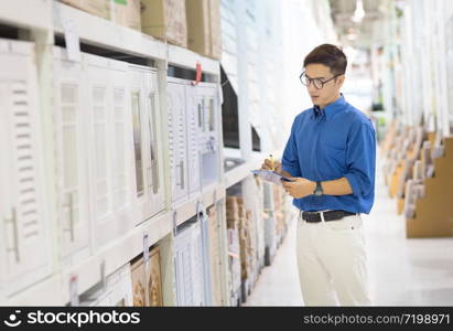 Asian manager man doing stocktaking of products management in cardboard box on shelves in warehouse using clipboard file and pen. Male professional assistant checking stock in factory.