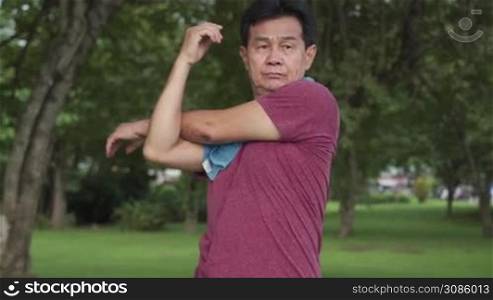 Asian mature man doing arm shoulder muscle stretching before exercise Inside public park with trees and green grass, morning exercising routine, active Motivation after retirement life, body condition