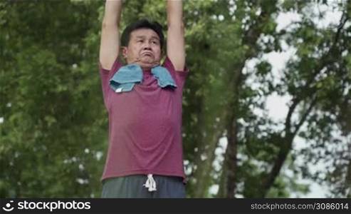 Asian mature man doing arm stretching warm up before exercise in the outdoor park with trees background, after morning exercise routine, active Motivation after retirement life, health care insurance