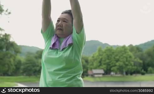 Asian mature woman doing over head arm stretching before exercise in the outdoor park with trees mountain background, morning exercise routine, active elderly aged woman, retirement life stronger body