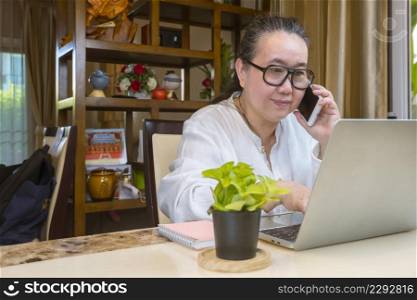 Asian mature woman talking on smartphone and using laptop computer while work from home in living room
