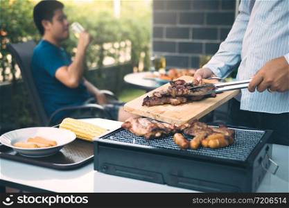 Asian men are pinching pork on a wooden cutting board and holding it to friends who are celebrating in the back.