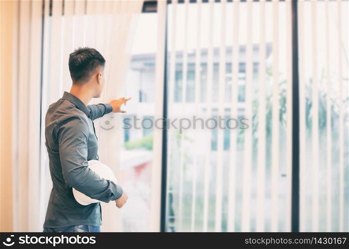 Asian men engineering holding white hard hat for working at office and open the curtains for looking construction