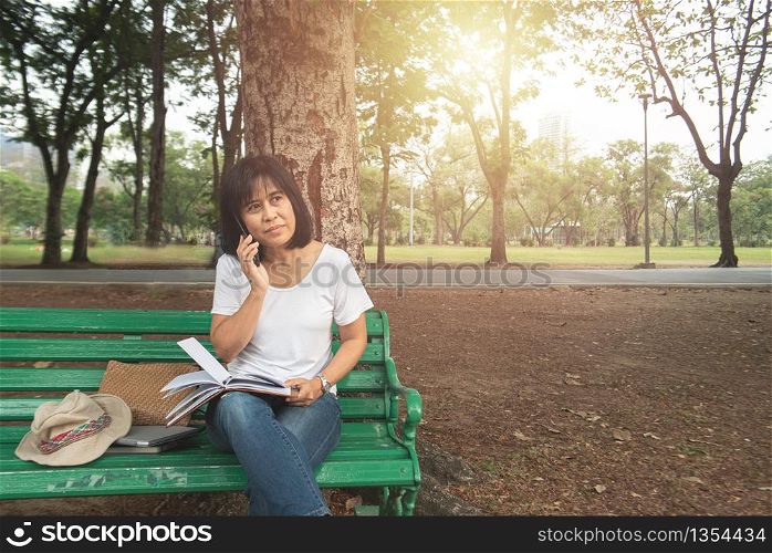 Asian mid age happy woman using smart mobile phone dress casual sitting outside in nature spring park.