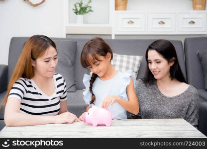 Asian mother and aunt smile and happy see daughter putting coin in piggy bank for saving in the living room at home, kid put money with deposit banking for future, family concept.