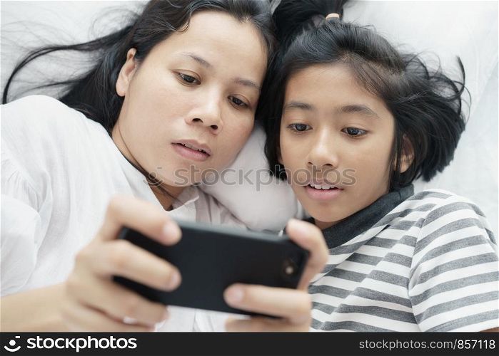 Asian mother and daughter playing game with phone together on the bed. Women and girl so happy and fun in bedroom.