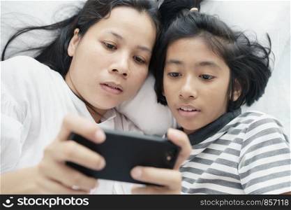 Asian mother and daughter playing game with phone together on the bed. Women and girl so happy and fun in bedroom.