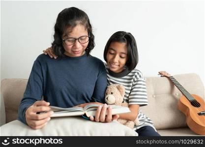 Asian mother and daughter read a book while sitting on the sofa. The little girl smiled and hugged his doll in the living room at home.