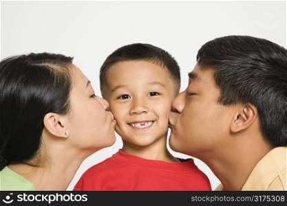 Asian mother and father kissing opposite cheeks of smiling son in front of white background.