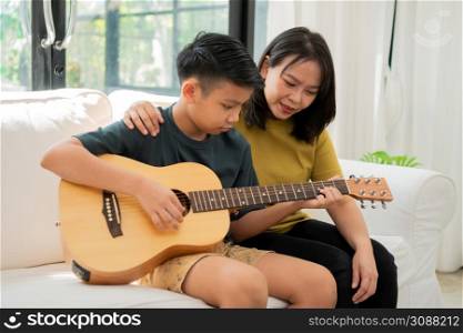 Asian mother embraces son, Asian boy playing guitar and mother embrace on the sofa and feel appreciated and encouraged. Concept of a happy family, learning and fun lifestyle, love family ties