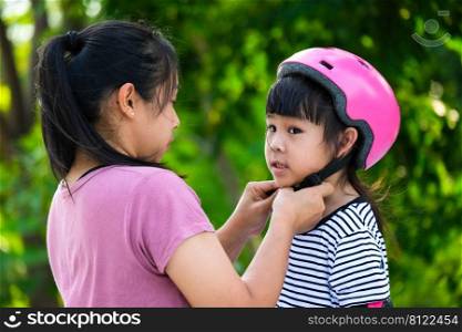 Asian mother helps daughter put on protective pads and safety helmet before practicing roller skating in the park. Exciting outdoor activities for kids.
