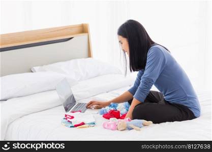 Asian mother shopping online for her baby clothes on laptop computer and tablet on the bed she makes purchase new baby clothes for an unborn baby preparing for the new child