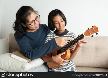 Asian mother teaches her daughter to play ukulele while sitting on the sofa in her home living room.