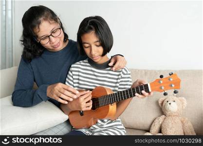 Asian mother teaches her daughter to play ukulele while sitting on the sofa in her home living room.