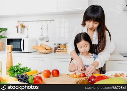 Asian Mother teaching her little child using knife to slice apple for cook food in the kitchen.