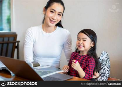 Asian mothers are teaching their daughters to read a book and use notebooks and technology for online learning during school holidays at home. Educational concepts and activities of the family