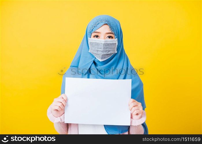 Asian Muslim Arab, Portrait of beautiful young woman religious wear veil hijab and face mask protective to prevent coronavirus she hold white paper blank board in chest, isolated on yellow background