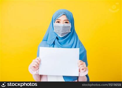 Asian Muslim Arab, Portrait of beautiful young woman religious wear veil hijab and face mask protective to prevent coronavirus she hold white paper blank board in chest, isolated on yellow background