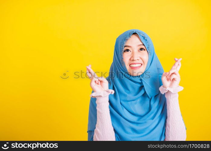 Asian Muslim Arab, Portrait of happy Asian beautiful young woman Islamic religious wear veil hijab she smiling and holding fingers crossed for good luck, studio shot isolated on yellow background