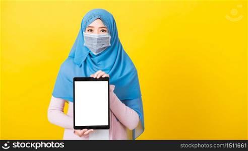Asian Muslim Arab, Portrait of happy beautiful young woman Islam religious wear veil hijab and face mask protect quarantines disease coronavirus show blank screen tablet isolated yellow background