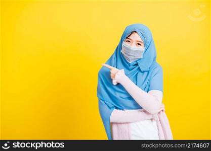 Asian Muslim Arab, Portrait of happy beautiful young woman religious wear veil hijab and face mask protective to prevent coronavirus she pointing finger side away space, isolated on yellow background