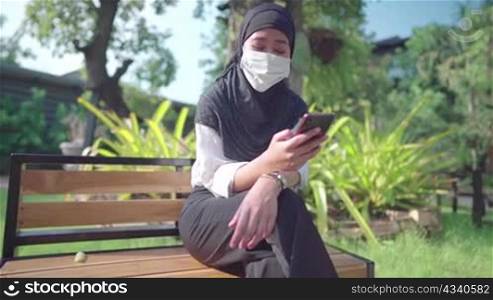 Asian Muslim female wear hijab and protective face mask. using smartphone sitting down relaxing time inside public park, New normal COVID pandemic, outdoors sunny day, wireless technology connection